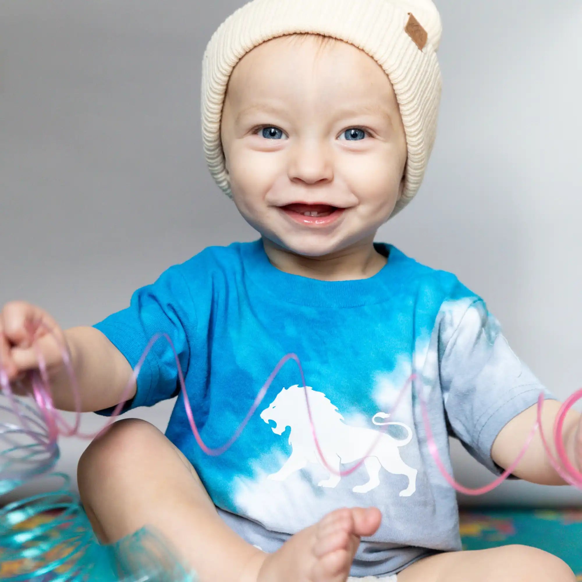 A smiling toddler wearing a cream beanie and blue dinosaur t-shirt.